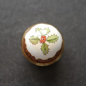 以下が含まれることがあります： ゴールドの金属製ベースと白いセラミック製のトップを備えた丸いキャビネットノブ。トップには、白い背景の中央に緑のヒイラギの葉と赤い実が手描きで描かれており、伝統的なクリスマスプディングのようなデザインになっています。