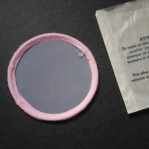 May include: A circular sifter featuring a fine grey mesh screen surrounded by a soft pink fabric border. Next to the sifter is a translucent paper packet with printed text that reads: This sifter is for compact use.