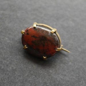 May include: An oval-shaped brooch featuring a polished, dark reddish-brown stone with black mottled inclusions. The stone is secured in a gold-toned metal setting with six small prongs holding the edges, and a thin metal pin extends from the side.