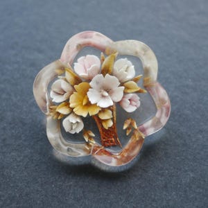 May include: A clear, flower-shaped brooch featuring a bouquet of white, pink, and yellow flowers. The brooch has a clear, petal-shaped border with a pinkish tint. The flowers are arranged in a vase with brown and gold detailing.
