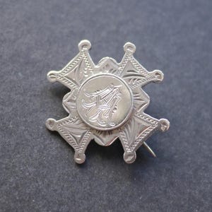 May include: A silver-toned brooch with an intricate starburst design. The brooch features a central circular emblem with an ornate letter "A" and is accented with detailed patterns and a pin backing.