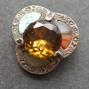 May include: A silver-toned brooch featuring an ornate, floral-engraved metal frame. The centre holds a large, round, faceted amber-coloured stone. Three smaller, crescent-shaped cabochon stones in pale blue, white with orange banding, and muted green surround it.