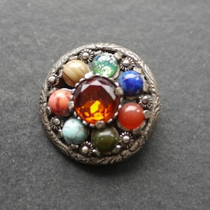May include: A vintage silver brooch with a central amber stone and an array of colourful gemstones. The brooch has a floral border and features stones in red, blue, green, and turquoise. The brooch is approximately 3 cm in diameter.