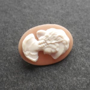 May include: An oval-shaped cameo brooch featuring a white relief carving of a profile portrait of a person with wavy hair set against a smooth, light peach-coloured background. The carving displays intricate details of the facial features and hair.