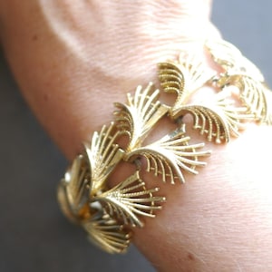 May include: A gold-tone metal link bracelet featuring a repeating pattern of stylised, fan-shaped leaf motifs. Each individual link is connected to form a flexible band that wraps around the wrist, showcasing a textured, metallic finish throughout.