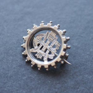 May include: A silver-colored brooch with a circular design. The brooch features a gear-like outer ring with a detailed, ornate emblem in the center. The pin is set against a dark gray background.