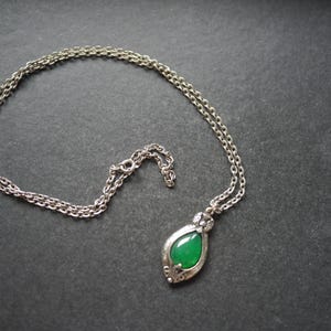 May include: A silver-toned metal chain necklace featuring a teardrop-shaped pendant. The pendant contains a smooth, polished green stone set within an ornate, textured silver-coloured metal frame that tapers to a point at the bottom.
