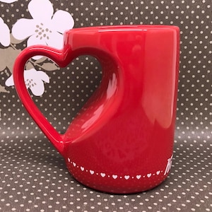 May include: A bright red ceramic mug featuring a distinct heart-shaped handle. The base of the mug is decorated with a horizontal band of small white hearts, and a partial white script text is visible on the lower right side of the exterior.