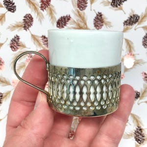 May include: A small white ceramic cup held within a decorative silver-toned metal holder. The holder features an intricate, repeating lattice pattern with a thin, curved metal handle attached to the side, providing a secure grip for the cup.