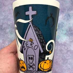 May include: A white ceramic tumbler with a Halloween-themed design. The design features a purple tombstone with the word "ZERO" and a ghost, bats, and jack-o'-lanterns. The tumbler has a dark teal background and a white rim.