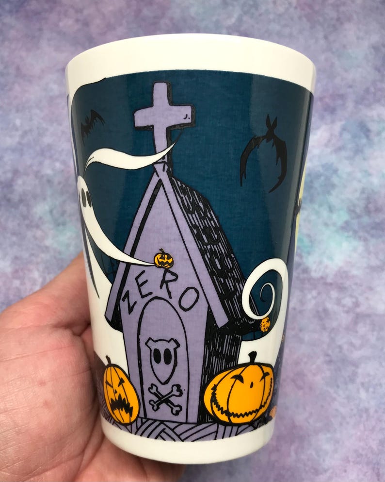 May include: A white ceramic tumbler with a Halloween-themed design. The design features a purple tombstone with the word "ZERO" and a ghost, bats, and jack-o'-lanterns. The tumbler has a dark teal background and a white rim.