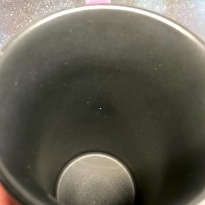 May include: A black travel mug with a pink handle. The interior of the mug is black, and the bottom is a lighter shade of gray. The mug is cylindrical and appears to be made of metal.