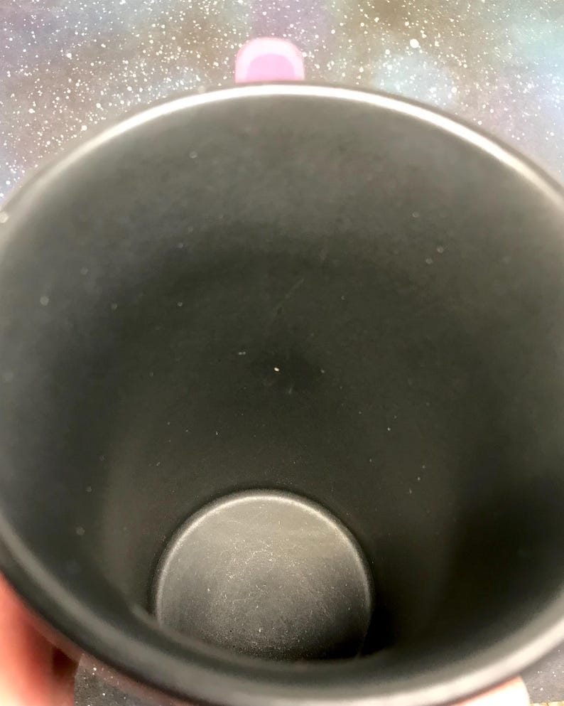 May include: A black travel mug with a pink handle. The interior of the mug is black, and the bottom is a lighter shade of gray. The mug is cylindrical and appears to be made of metal.