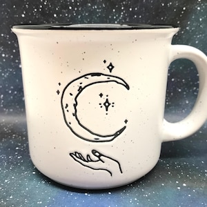 May include: A white ceramic mug with a speckled finish and a black rim. The front features a black line art illustration of a crescent moon surrounded by small stars, positioned directly above an open hand reaching upward toward the moon.