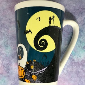 May include: Tall white ceramic mug featuring a design from The Nightmare Before Christmas. The mug has a dark teal background with a yellow full moon, Jack and Sally silhouettes, and swirling hill. Orange jack-o'-lanterns and bats are also featured.
