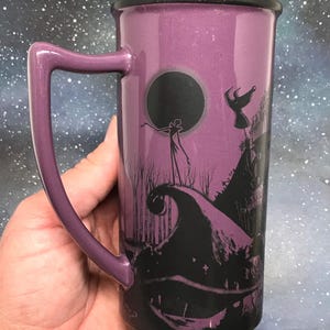 May include: Purple ceramic mug featuring a black silhouette design from The Nightmare Before Christmas. The mug has a black rim and handle, and the design includes Jack Skellington and other characters. The mug is held in a hand.