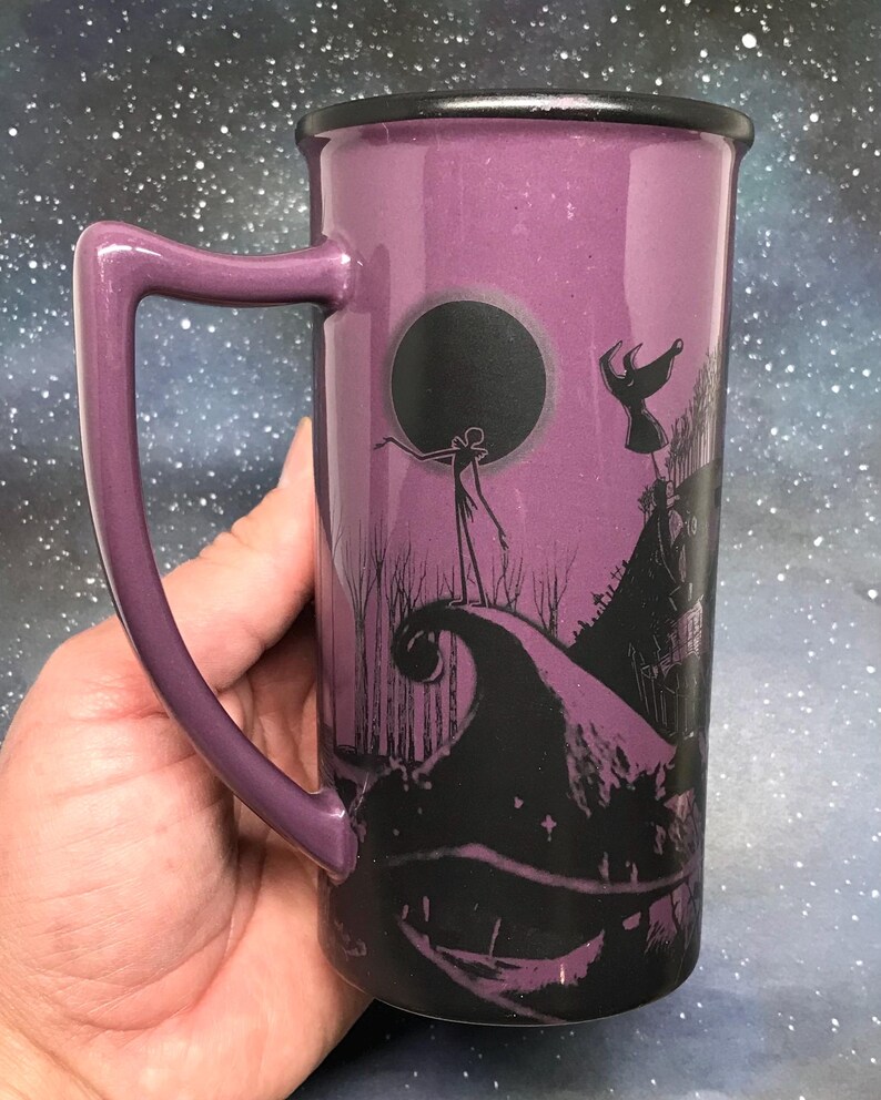 May include: Purple ceramic mug featuring a black silhouette design from The Nightmare Before Christmas. The mug has a black rim and handle, and the design includes Jack Skellington and other characters. The mug is held in a hand.
