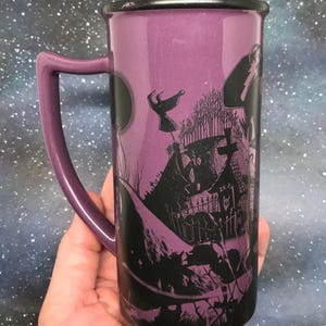 May include: Purple ceramic mug with a black rim and handle. The mug features a black and white illustration of a spooky graveyard scene with a full moon and flying figures. The handle is a matching purple color.