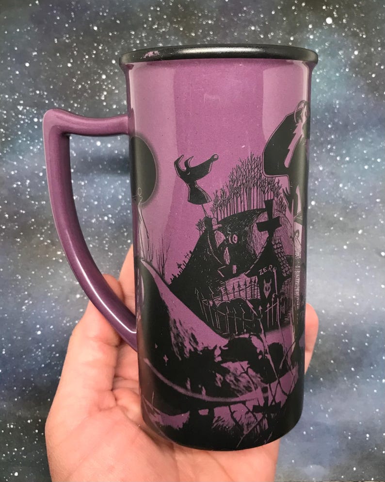 May include: Purple ceramic mug with a black rim and handle. The mug features a black and white illustration of a spooky graveyard scene with a full moon and flying figures. The handle is a matching purple color.