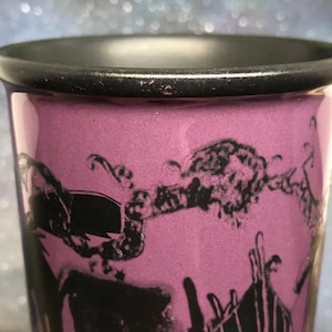 May include: Close-up of a purple shot glass with a black rim and a black graphic design. The design appears to be a stylized illustration. The glass has a smooth, glossy finish.
