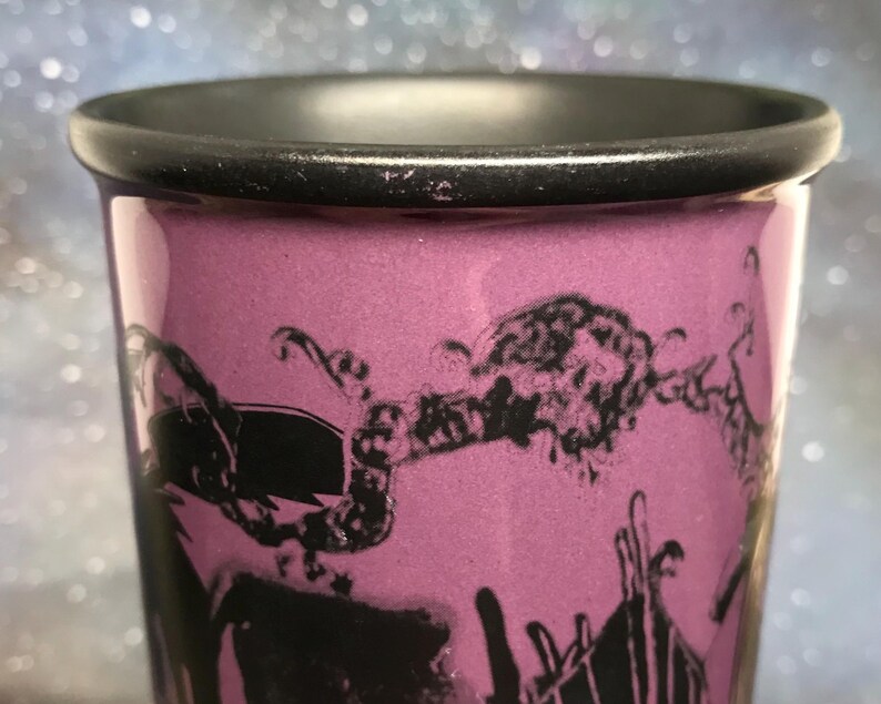 May include: Close-up of a purple shot glass with a black rim and a black graphic design. The design appears to be a stylized illustration. The glass has a smooth, glossy finish.