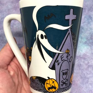 May include: White ceramic mug featuring Zero from The Nightmare Before Christmas. The design includes Zero, a tombstone with "ZERO" on it, bats, and jack-o'-lanterns. The mug has a white handle and a dark teal background.