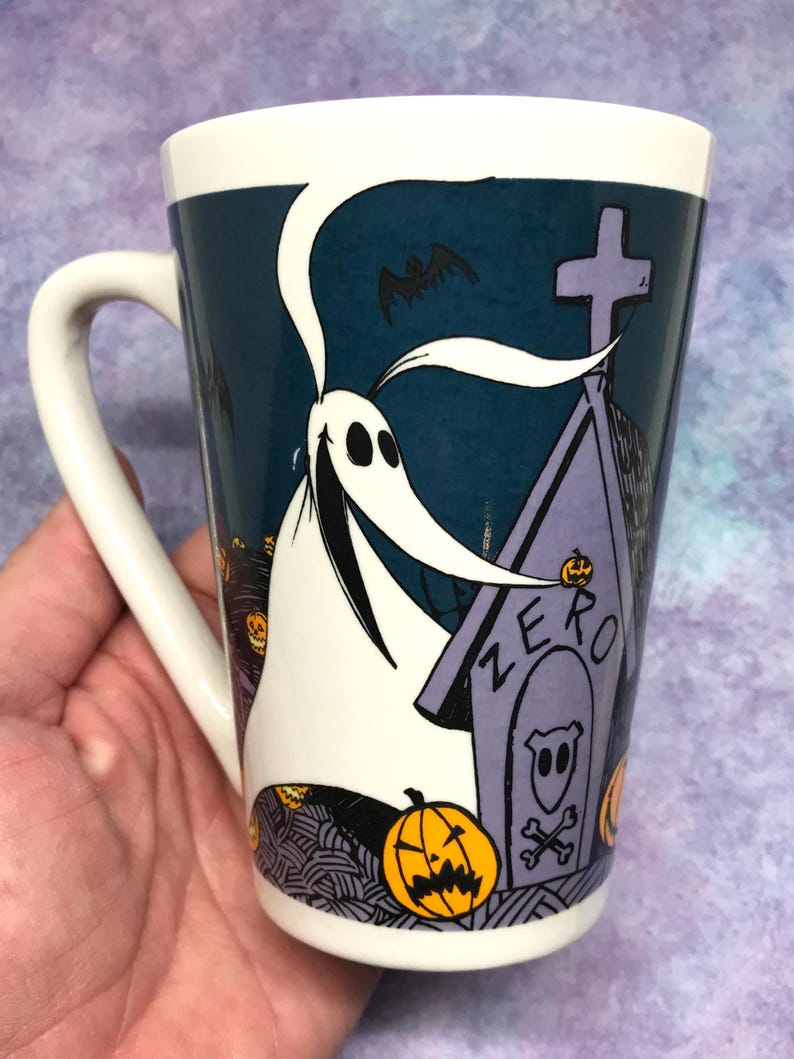 May include: White ceramic mug featuring Zero from The Nightmare Before Christmas. The design includes Zero, a tombstone with "ZERO" on it, bats, and jack-o'-lanterns. The mug has a white handle and a dark teal background.