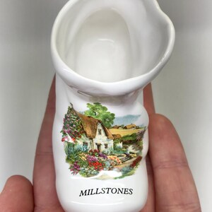 May include: White ceramic boot-shaped vase with a scenic illustration of a cottage, trees, and flowers. The vase has the word "MILLSTONES" printed on the front. The vase is held in a person's hand.