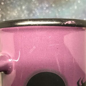 May include: Close-up of a pink enamel mug with a black rim and handle. The mug has a black graphic design at the bottom. The mug is a light pink color.