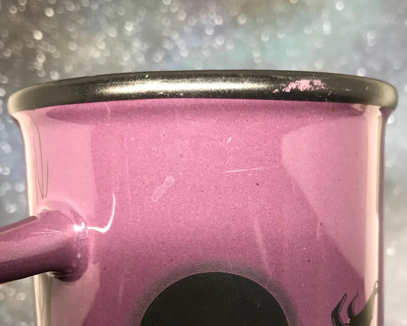 May include: Close-up of a pink enamel mug with a black rim and handle. The mug has a black graphic design at the bottom. The mug is a light pink color.