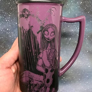 May include: Purple ceramic mug featuring Sally from The Nightmare Before Christmas. The mug has a black rim and handle, with a detailed illustration of Sally in a patchwork dress. The background includes a spooky silhouette of a graveyard.