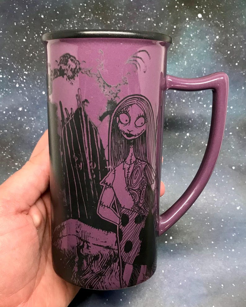 May include: Purple ceramic mug featuring Sally from The Nightmare Before Christmas. The mug has a black rim and handle, with a detailed illustration of Sally in a patchwork dress. The background includes a spooky silhouette of a graveyard.
