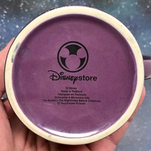 May include: Purple ceramic mug with a white rim and handle. The bottom of the mug features the Disney Store logo and text indicating it is from the movie "Tim Burton's The Nightmare Before Christmas."