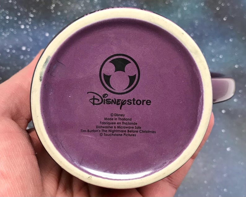May include: Purple ceramic mug with a white rim and handle. The bottom of the mug features the Disney Store logo and text indicating it is from the movie "Tim Burton's The Nightmare Before Christmas."