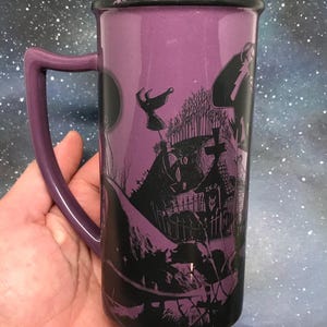 May include: A tall, purple ceramic mug with a black rim and handle. The mug features a black and white illustration of a spooky graveyard scene with a full moon. The handle is a matching purple color. The mug is held in a hand.