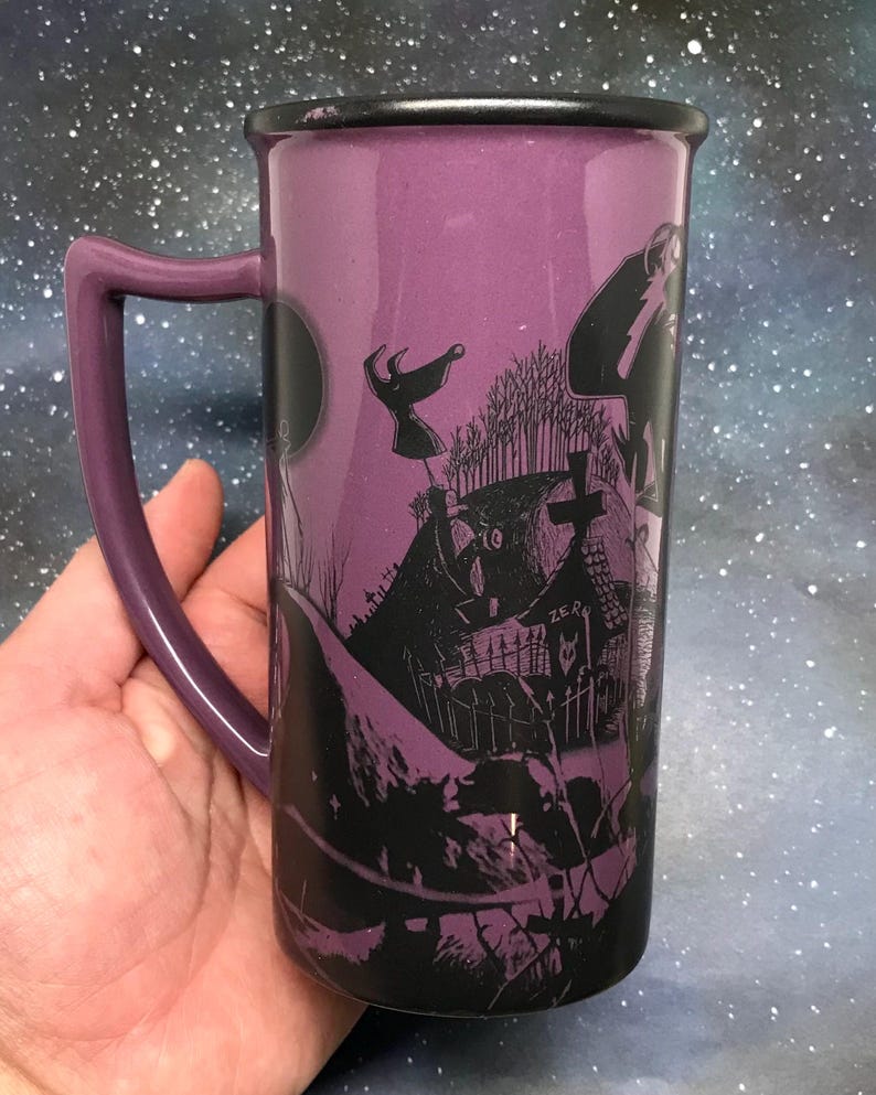 May include: A tall, purple ceramic mug with a black rim and handle. The mug features a black and white illustration of a spooky graveyard scene with a full moon. The handle is a matching purple color. The mug is held in a hand.