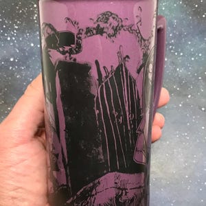 May include: Purple ceramic mug with a black rim and handle. The mug features a black and purple graphic design of a spooky scene with a gate and characters. The mug is held in a hand.