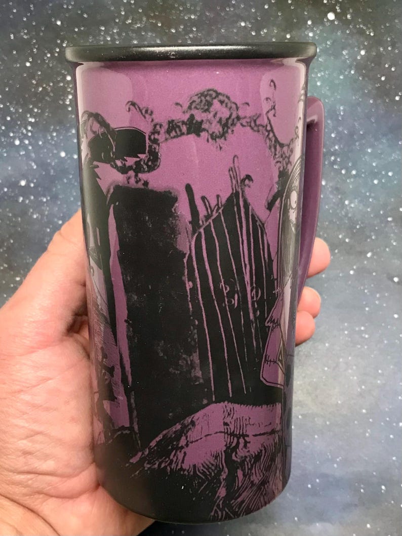 May include: Purple ceramic mug with a black rim and handle. The mug features a black and purple graphic design of a spooky scene with a gate and characters. The mug is held in a hand.