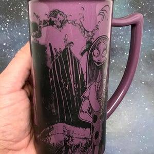 May include: A tall, purple ceramic mug featuring a black and white illustration of Sally from The Nightmare Before Christmas. The mug has a matching purple handle and a black rim. The background is a starry night sky.