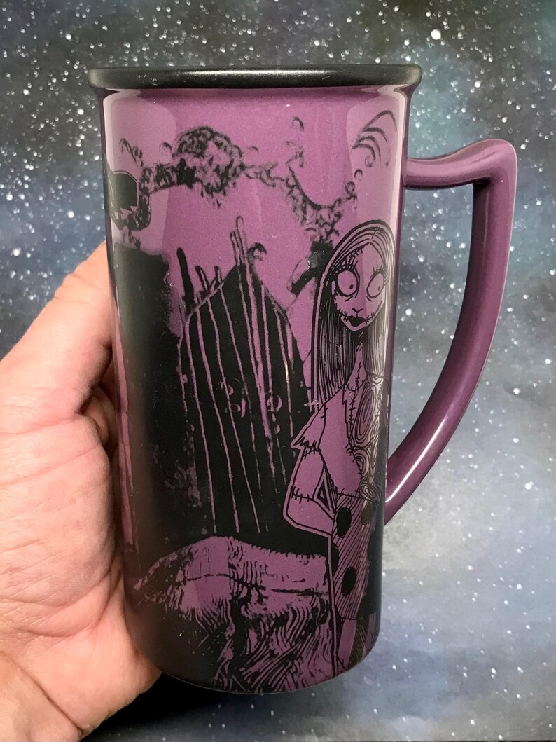 May include: A tall, purple ceramic mug featuring a black and white illustration of Sally from The Nightmare Before Christmas. The mug has a matching purple handle and a black rim. The background is a starry night sky.