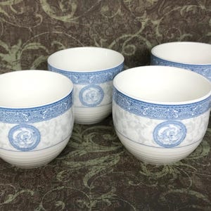 May include: A set of four white ceramic tea cups featuring a blue and white decorative band around the rim and a circular blue floral motif on the front. The cups have a smooth, rounded shape with a subtle ribbed texture near the base.