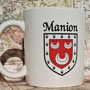 May include: White ceramic mug featuring the name "Manion" in black script above a family crest. The crest is red with three white crescent moon shapes and black and white decorative elements. The mug has a curved handle.