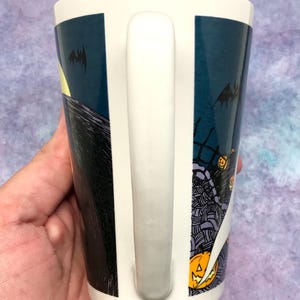 May include: White ceramic mug with a wraparound design featuring a spooky Halloween scene. The design includes a full moon, bats, and a graveyard with jack-o'-lanterns. The mug has a white handle.