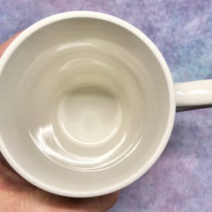 May include: A white ceramic mug with a rounded handle. The mug is empty and viewed from above, showing the interior and the handle on the right side. The mug is held by a hand.