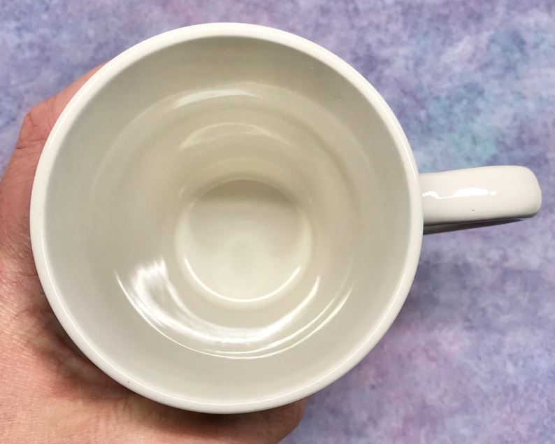 May include: A white ceramic mug with a rounded handle. The mug is empty and viewed from above, showing the interior and the handle on the right side. The mug is held by a hand.