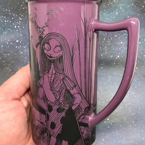 May include: Purple ceramic mug featuring a black and white illustration of Sally from The Nightmare Before Christmas. The mug has a matching purple handle and a black rim. The design includes trees and a spooky background.