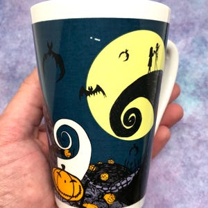 May include: A ceramic mug featuring a design from "The Nightmare Before Christmas." The mug has a white handle and rim, with a dark blue background. The design includes a yellow moon, bats, and Jack and Sally silhouettes, with pumpkins and swirls.