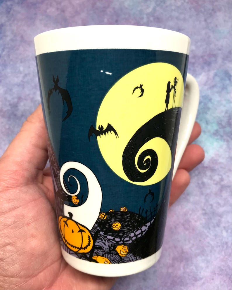 May include: A ceramic mug featuring a design from "The Nightmare Before Christmas." The mug has a white handle and rim, with a dark blue background. The design includes a yellow moon, bats, and Jack and Sally silhouettes, with pumpkins and swirls.