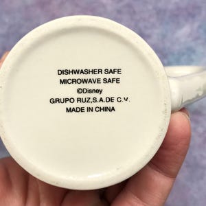 May include: A white ceramic mug with a handle, featuring text that reads "Dishwasher Safe", "Microwave Safe", the Disney logo, and "Made in China". The mug is held in a hand.