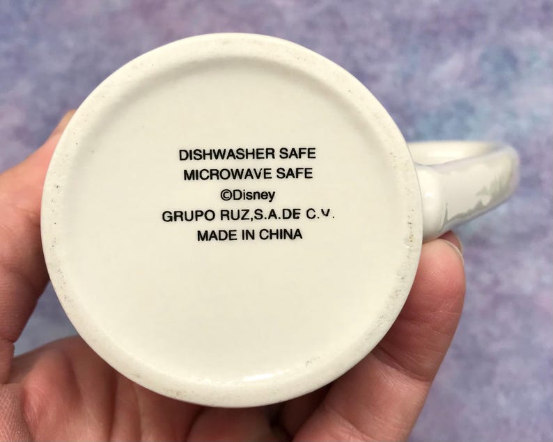 May include: A white ceramic mug with a handle, featuring text that reads "Dishwasher Safe", "Microwave Safe", the Disney logo, and "Made in China". The mug is held in a hand.